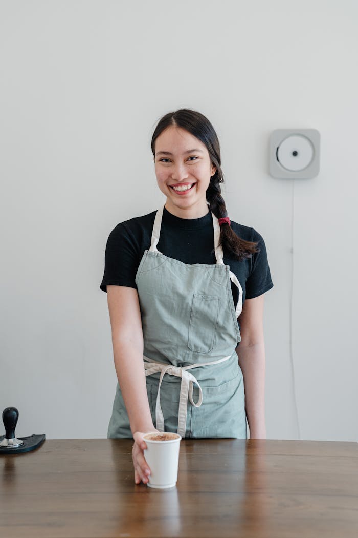 Positive young Asian woman wearing apron giving paper cup with coffee to go while standing at wooden counter during work in modern coffee house and looking at camera with smile