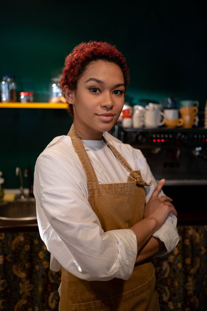 A smiling barista stands confidently in a cozy café, ready to serve with a warm demeanor.